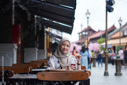 Indonesian Hijabi Woman Smiling with Refreshment at Cafe Bench. Joyful Moments at the cafe concept. photo