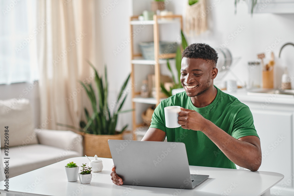 Smiling black man enjoying cup of coffee while using laptop in kitchen, freelancer at home Smiling black man enjoying cup of coffee while using laptop in kitchen, freelancer at home
