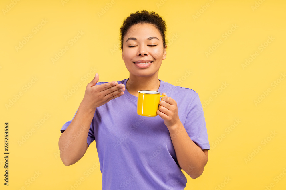Smiling African American woman wearing t shirt holding yellow cup with closed eyes sniffing coffee Smiling African American woman wearing t shirt holding yellow cup with closed eyes sniffing coffee