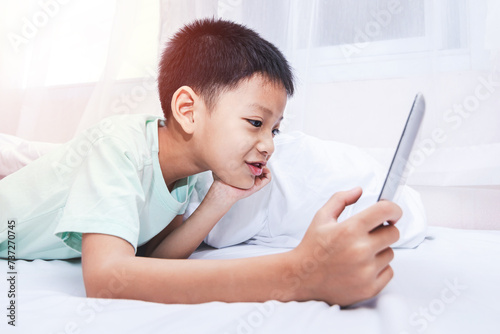 little boy looking at a tablet and playing games while lying on the bed at home. kid using smart devices online with his friends in a video call.
