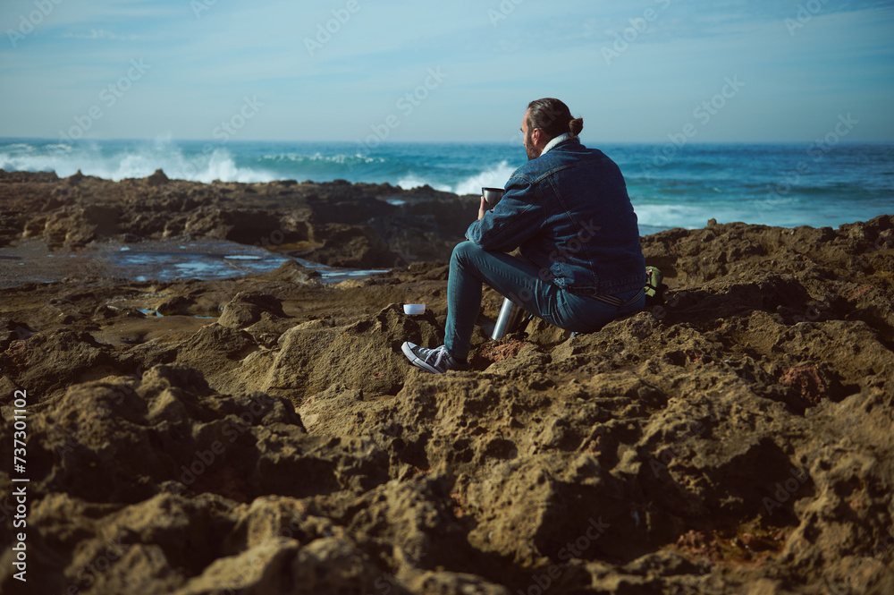 Mature Caucasian bearded man taking coffee break while hiking in the rocky beach, sitting relaxed on the rocky cliff