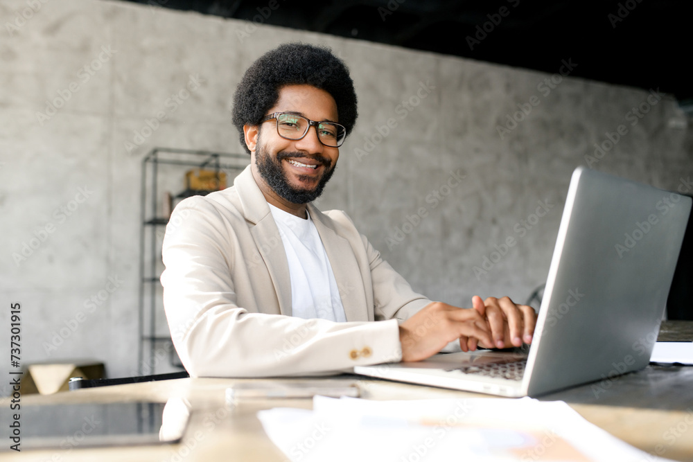 Cheerful young professional with trendy afro hairstyle confidently uses his laptop in a modern workspace, his smart casual attire perfectly blending with the innovative and stylish office environment