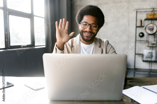 The young businessman in a modern workspace engages in an online conversation, with a friendly wave that showcases his personable nature and adaptability in a digital era. Video call concept