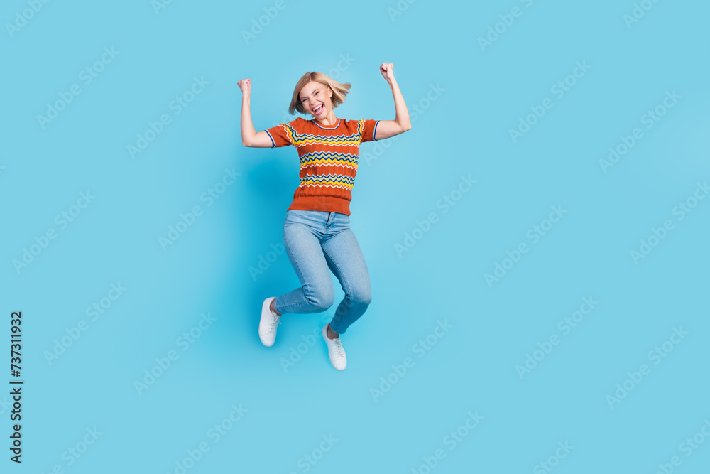 Full size photo of good mood overjoyed woman dressed knit t-shirt raising fists up jumping win bet isolated on blue color background Full size photo of good mood overjoyed woman dressed knit t-shirt raising fists up jumping win bet isolated on blue color background