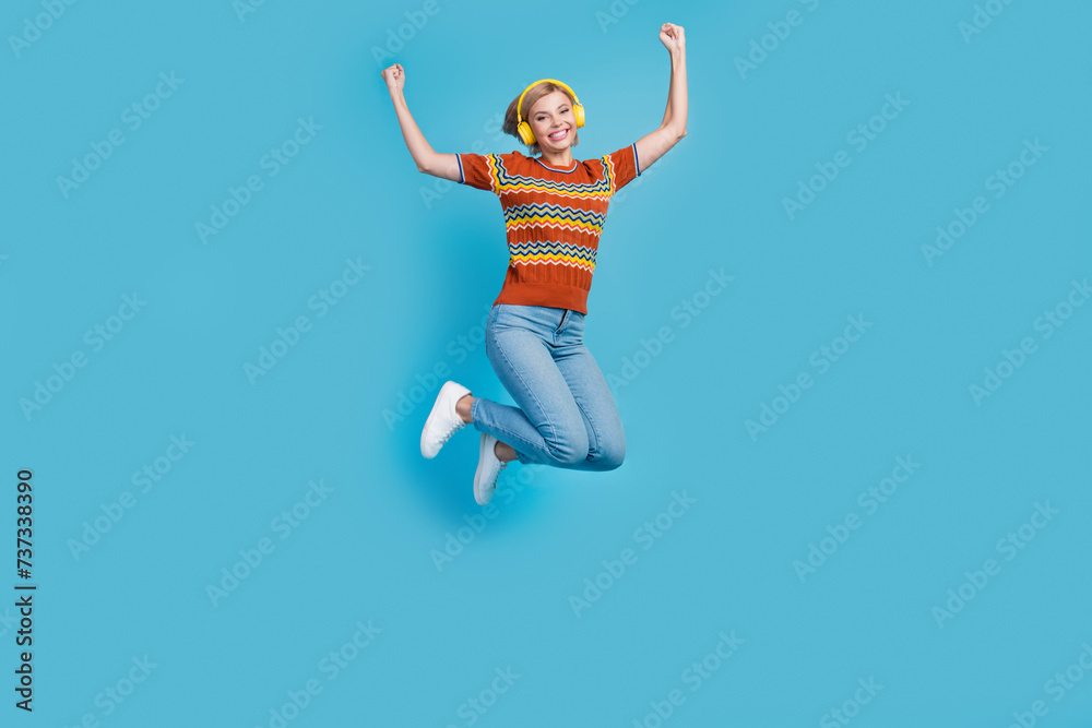 Full size photo of overjoyed woman dressed knit t-shirt denim trousers flying in headphones arms up isolated on blue color background Full size photo of overjoyed woman dressed knit t-shirt denim trousers flying in headphones arms up isolated on blue color background