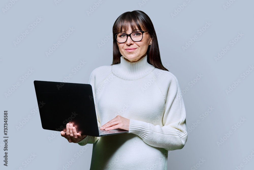 Middle-aged beautiful woman holding laptop looking at camera on grey background Middle-aged beautiful woman holding laptop looking at camera on grey background