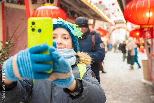 Happy 11 year old European child with a dragon mask on his hat takes a selfie at the Chinese New Year holiday fair, snowy winter.