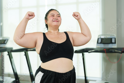 Plus-size Asian woman exercise in gym, Exuberant flexing muscles, showing strength and joy, treadmill machines in behind, flexing biceps, displaying strength and happiness in fitness setting