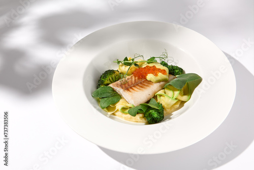 Cod fillet with caviar and zucchini-broccoli garnish, side view on white plate with delicate leaf shadow