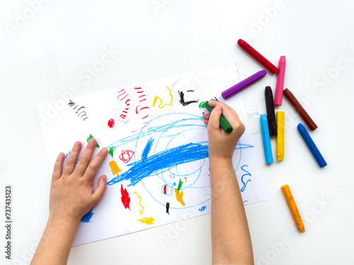 Childs hands drawing with colorful wax crayons on white paper, top view. Creative art concept for educational and developmental activities.