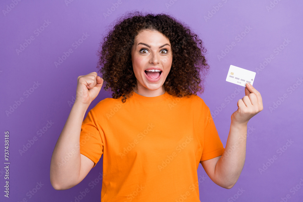 Photo of sweet lucky woman wear orange t-shirt rising fist holding credit card isolated purple color background Photo of sweet lucky woman wear orange t-shirt rising fist holding credit card isolated purple color background