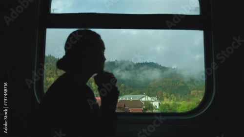Woman silhouette near the window in the train compartment. View on the mountains and small village through train window, fog over mountain peaks in the morning