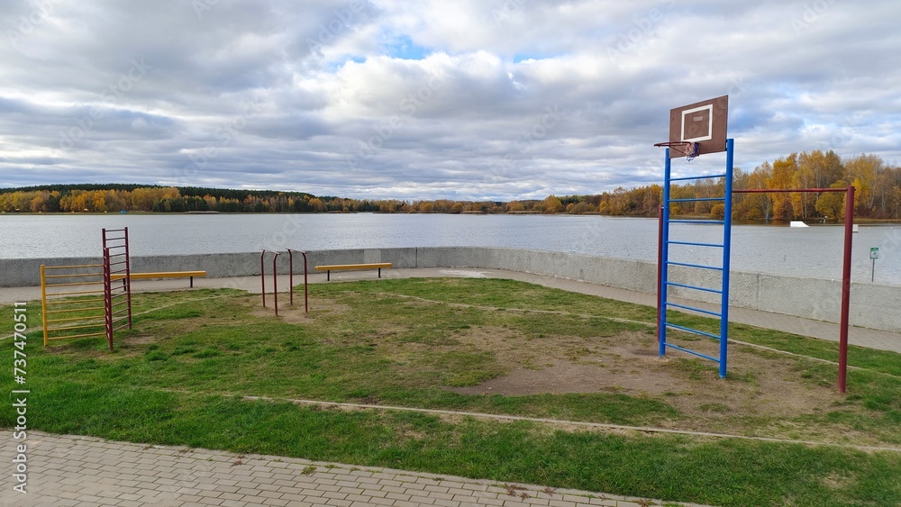 Near the concrete parapet on the lake shore, a plastic basketball backboard with a hoop is set up on a grassy lawn. Nearby there are tile paths and fitness equipment On the far shore there is a forest