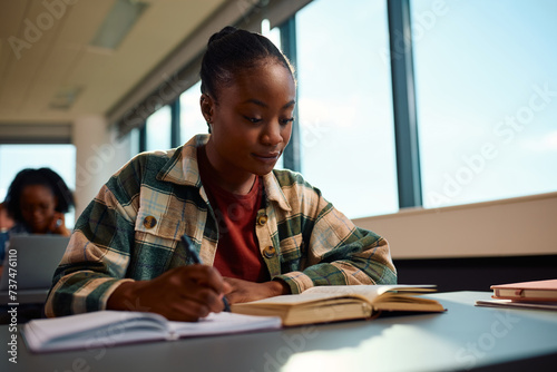 Black female student taking notes while learning at college classroom.