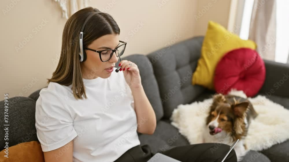 A focused hispanic woman works remotely with a headset in a cozy living room beside a resting sheltie dog. A focused hispanic woman works remotely with a headset in a cozy living room beside a resting sheltie dog.