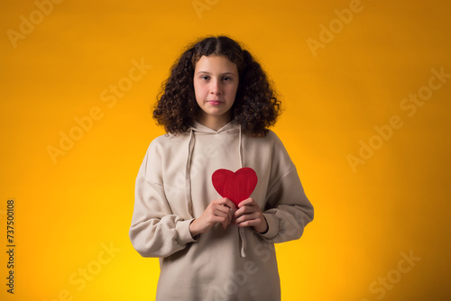 Smiling girl holding paper heart over yellow background. Love, care and Valentine day concept