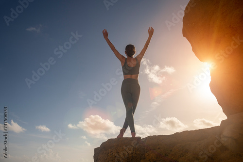 sporty woman standing on top of a rock at sunrise with her arms raised.