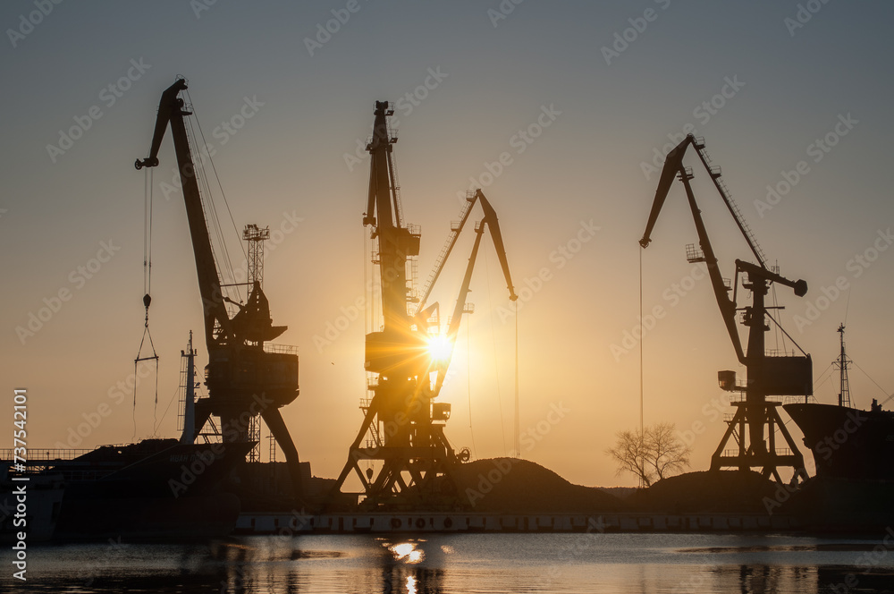 Golden sunrise illuminates industrial harbor cranes at port. Cargo for unloading, trade logistics. Heavy machinery silhouette against dawn sky, global shipping. Export, import at maritime terminal.