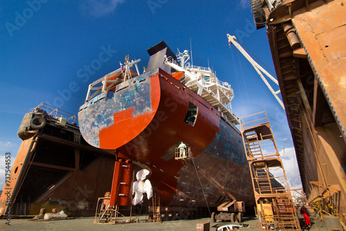 Commercial vessel undergoes maintenance in floating dock. Workers paint, repair hull, under clear skies at shipyard. Maritime industry, vessel refurbishment, drydock operation captured in daylight.