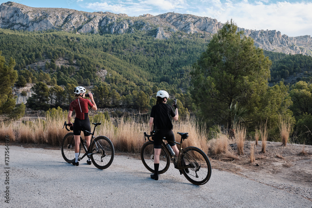 Fototapeta premium Two professional women cyclists riding gravel bicycles on a gravel road on a sunny summer day. Group bike ride in the natural countryside. Gravel advanture. Gravel cycling adventure. Alicante, Spain .