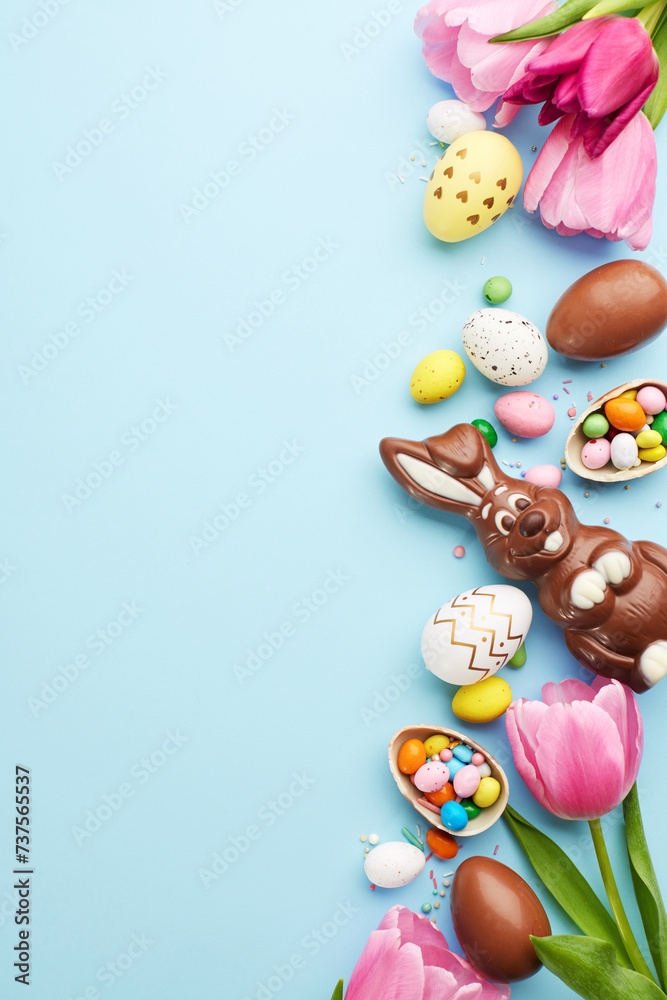 Easter joy: a bounty of sweets. Top view vertical shot of tulips and a delightful assortment of Easter chocolates and candies on a sky blue surface with space for heartfelt Easter wishes