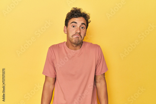 Young Latino man posing on yellow background shrugs shoulders and open eyes confused.