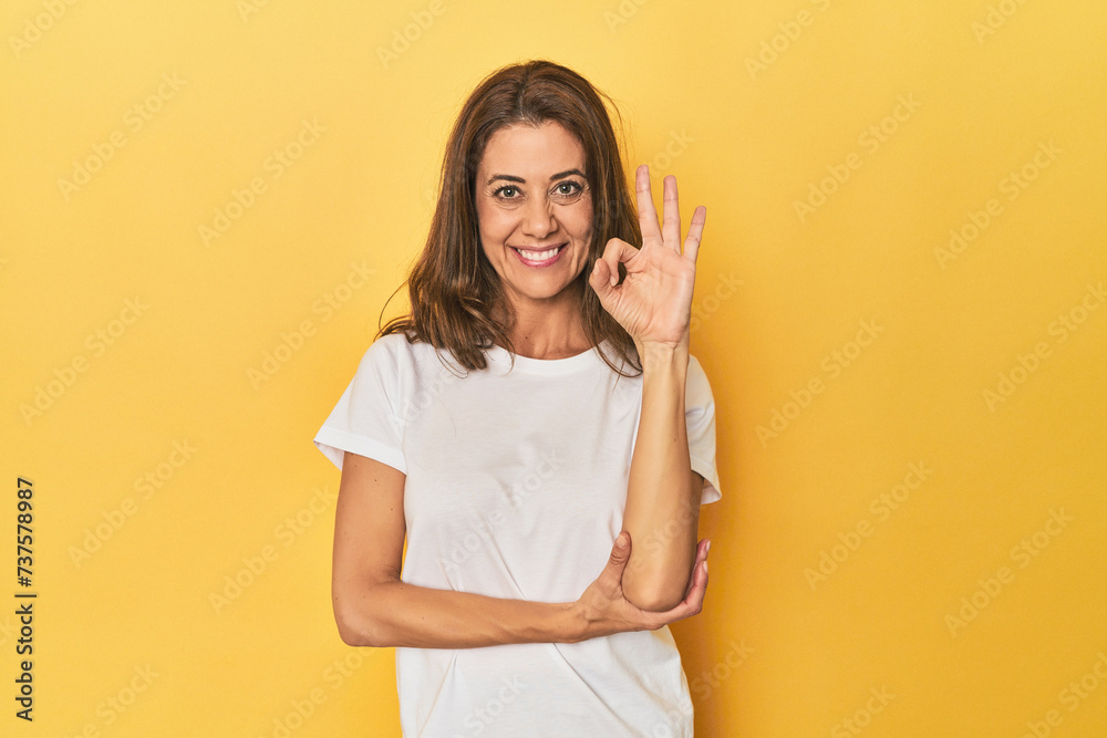 Middle-aged caucasian woman on yellow winks an eye and holds an okay gesture with hand. Middle-aged caucasian woman on yellow winks an eye and holds an okay gesture with hand.