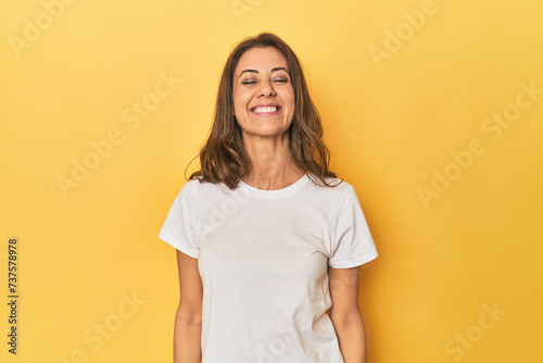 Middle-aged caucasian woman on yellow laughs and closes eyes, feels relaxed and happy.
