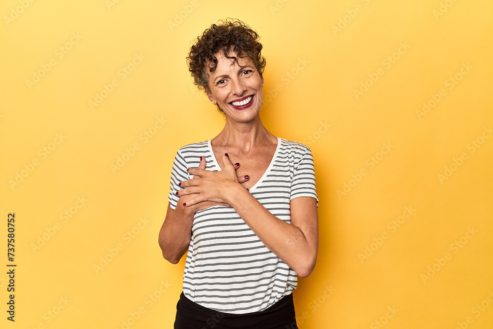 Mid-aged caucasian woman on vibrant yellow has friendly expression, pressing palm to chest. Love concept. Mid-aged caucasian woman on vibrant yellow has friendly expression, pressing palm to chest. Love concept.