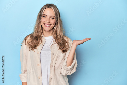 Blonde young caucasian woman in blue studio showing a copy space on a palm and holding another hand on waist. photo