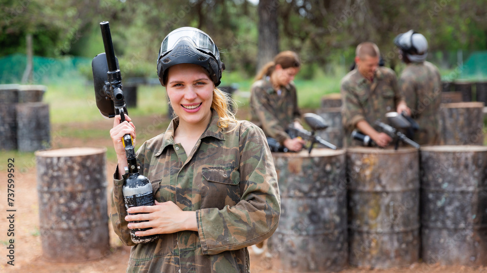 Fototapeta premium Young smiling woman paintball player in camouflage posing with gun outdoors