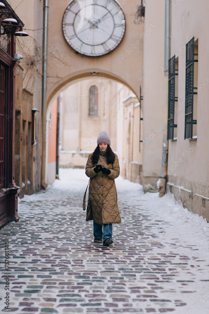 A young girl in a down jacket,a purple angora hat walks along a narrow old street with paving stones against the backdrop of a large wall clock,waiting for a date. Lviv,Armenian courtyard, Ukraine.
