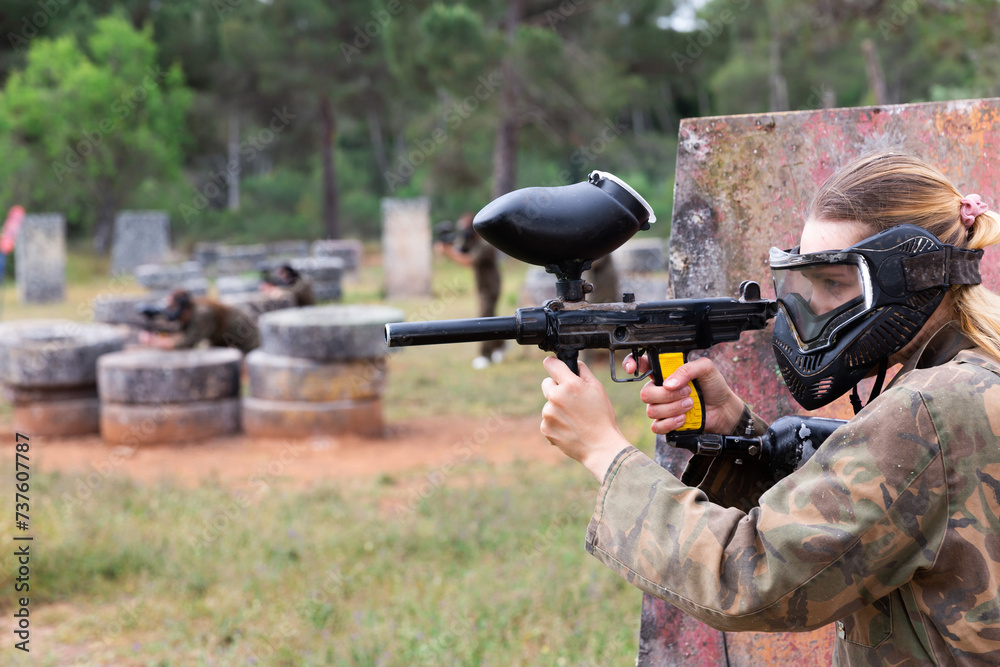 Fototapeta premium Portrait of girl paintball player in mask who is aiming in opponents on paintball field