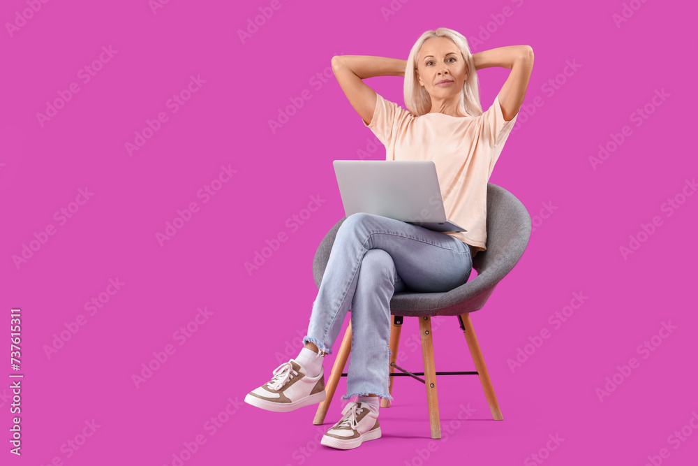 Thoughtful mature woman with laptop sitting in armchair on purple background Thoughtful mature woman with laptop sitting in armchair on purple background