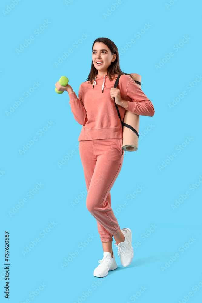 Beautiful young woman in stylish hoodie with dumbbell and yoga mat on blue background Beautiful young woman in stylish hoodie with dumbbell and yoga mat on blue background