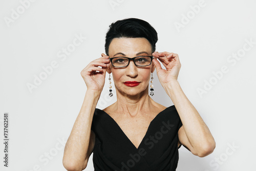 Elegant woman holding eyeglasses up to face in black dress with sophisticated look on white background
