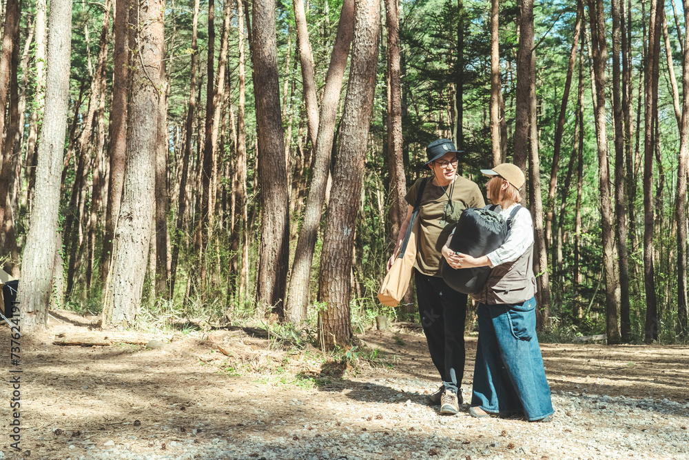 Obraz premium キャンプ場で寝袋を持って荷物を運ぶアジア人の夫婦キャンパー(グランピング・林間サイト)