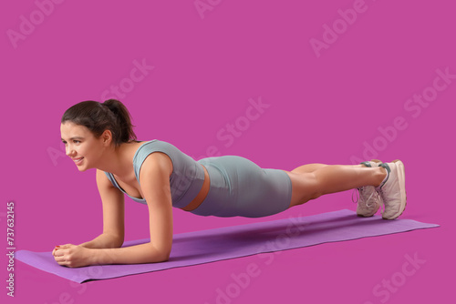 Fototapeta Naklejka Na Ścianę i Meble -  Young woman in sportswear doing plank on mat against purple background