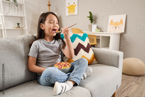 Cute little Asian girl eating cereal rings on sofa at home