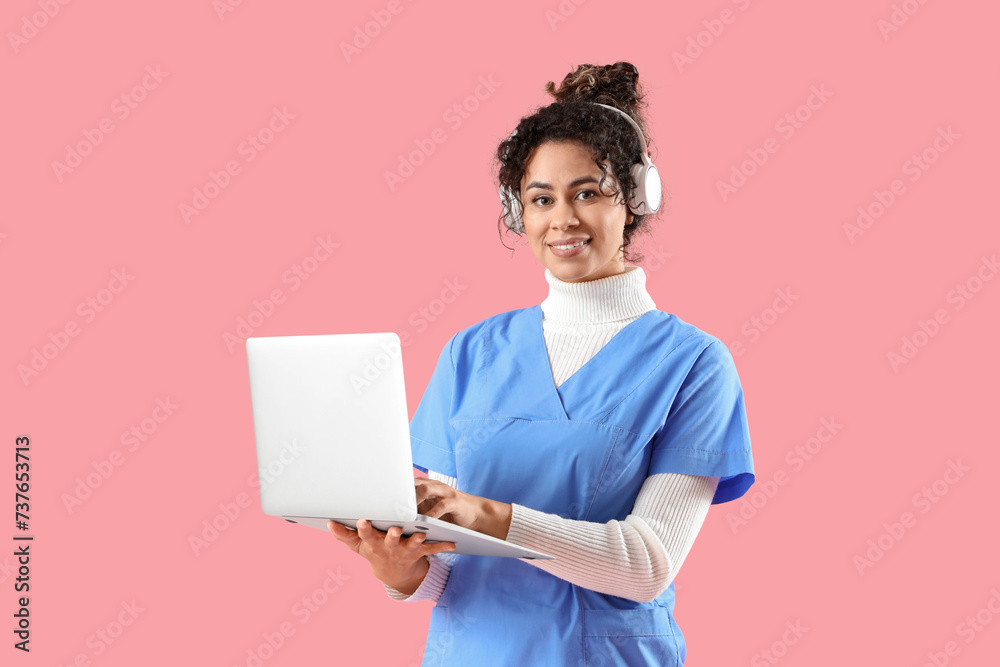 Female African-American medical intern in headphones using laptop on pink background