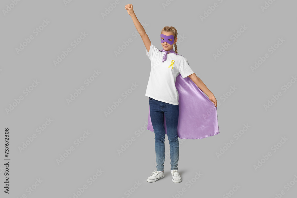 Cute teenage girl in superhero costume with yellow ribbon on grey background. International Childhood Cancer Day