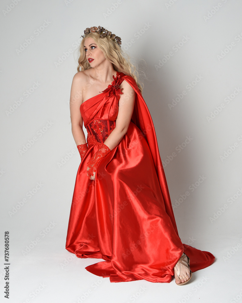 Full length portrait of beautiful blonde model dressed as ancient mythological fantasy goddess in flowing red silk toga gown, crown. Seated pose sitting on chair. isolated on white studio background. Full length portrait of beautiful blonde model dressed as ancient mythological fantasy goddess in flowing red silk toga gown, crown. Seated pose sitting on chair. isolated on white studio background.