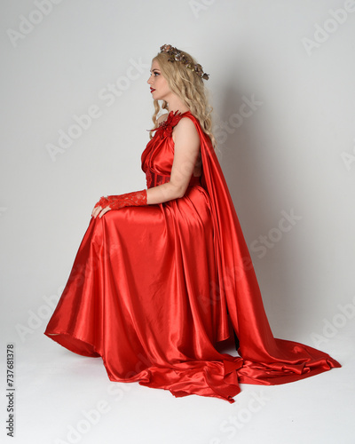 Full length portrait of beautiful blonde model dressed as ancient mythological fantasy goddess in flowing red silk toga gown, crown. Seated pose sitting on chair. isolated on white studio background.