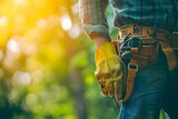 A laborer's hand in a used yellow glove stands out against a blurred, sunny green backdrop, with a tool belt around their waist