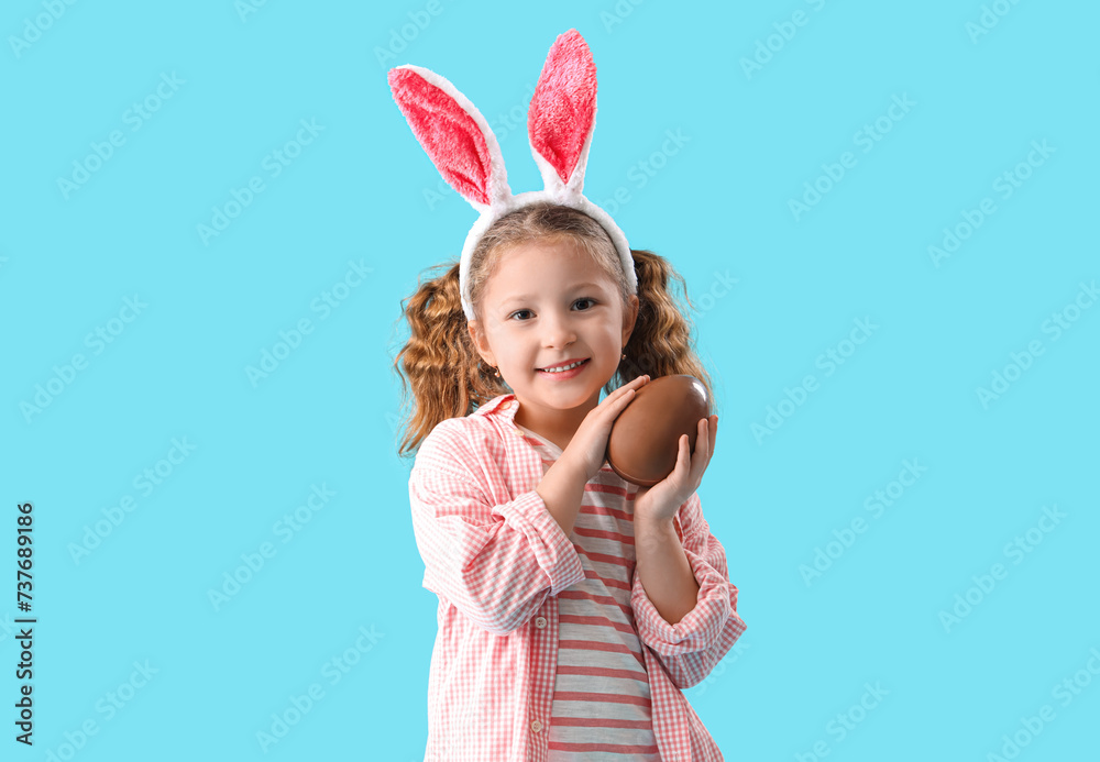 Cute little girl in bunny ears with chocolate Easter egg on blue background