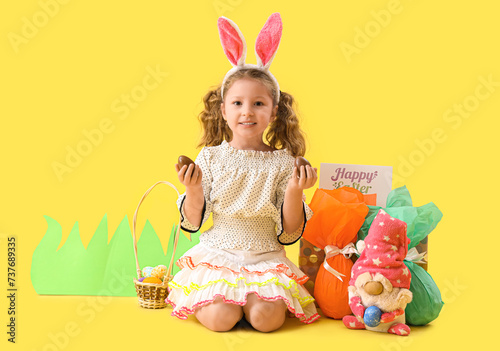Cute little girl in bunny ears with chocolate Easter eggs and decor sitting on yellow background