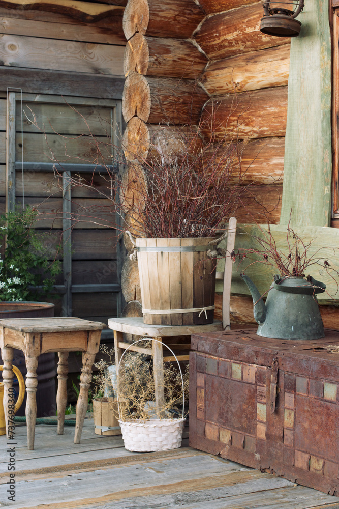 In the corner of a log house, a composition of old rustic items. A wooden bucket with branches, a green teapot, baskets with dry grass, a colored forged chest. Decoration. In the corner of a log house, a composition of old rustic items. A wooden bucket with branches, a green teapot, baskets with dry grass, a colored forged chest. Decoration.