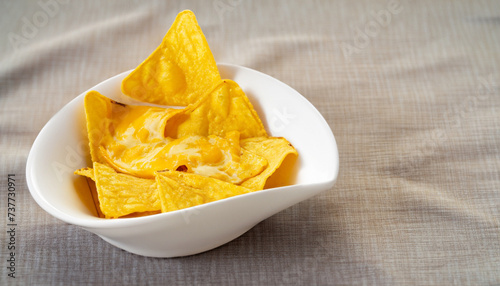 Single oval shaped white bowl of yellow tortilla chips topped with melted cheese placed on folded fabric napkin over tablecloth with copy space