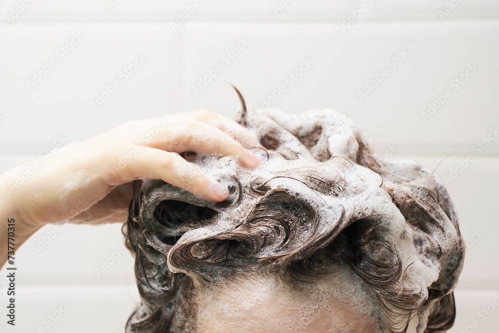A young woman washes her hair with shampoo on white tiles background.
