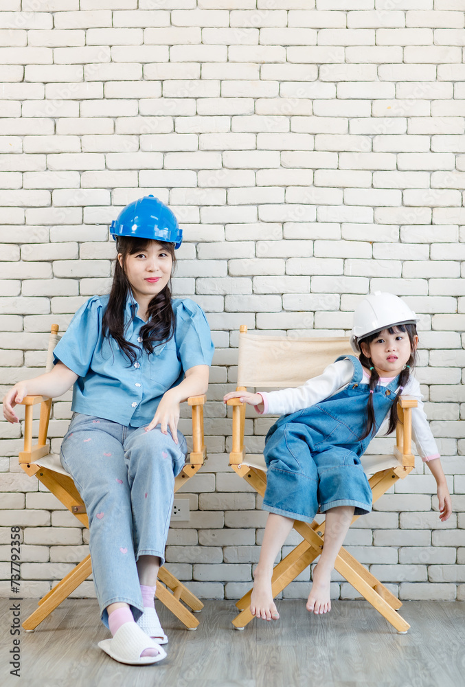 Asian beautiful young mother cute daughter little girl wearing engineer helmet sitting on a chair in white brick wall background studio portrait Mother's Day love family and Occupation kids concept. Asian beautiful young mother cute daughter little girl wearing engineer helmet sitting on a chair in white brick wall background studio portrait Mother's Day love family and Occupation kids concept.